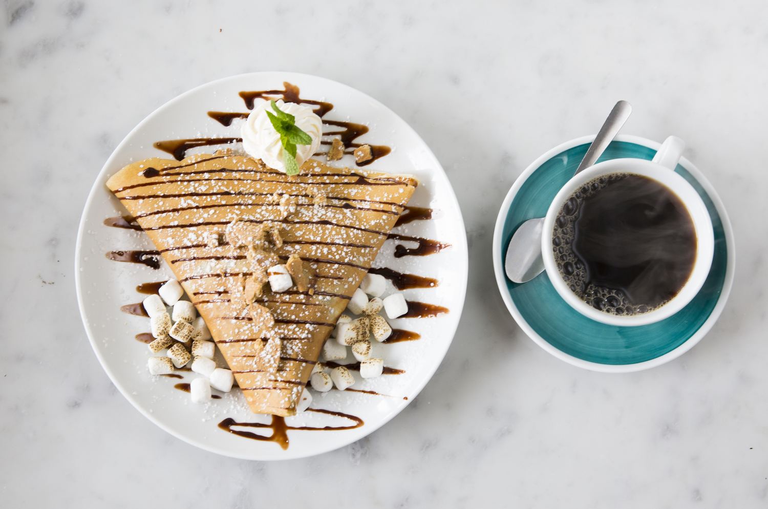 A S'mores crêpe drizzled with chocolate, toasted marshmallows and whipped cream next to a black coffee in a teal cup, on marble